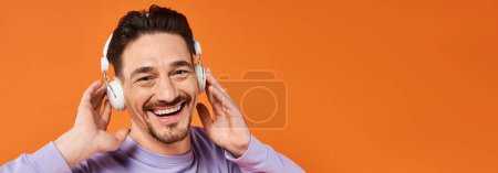 positiver Mann in lila Pullover und drahtlosen Kopfhörern, die Musik auf orangefarbenem Hintergrund hören, Banner