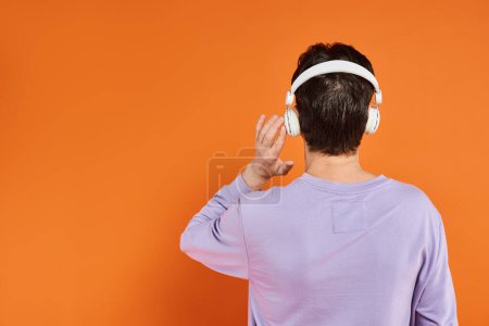 Rückansicht eines Mannes in lila Pullover und drahtlosen Kopfhörern, der Musik auf orangefarbenem Hintergrund hört