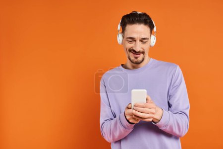 positiver Mann in drahtlosen Kopfhörern, der Musik hört und das Smartphone auf orangefarbenem Hintergrund benutzt