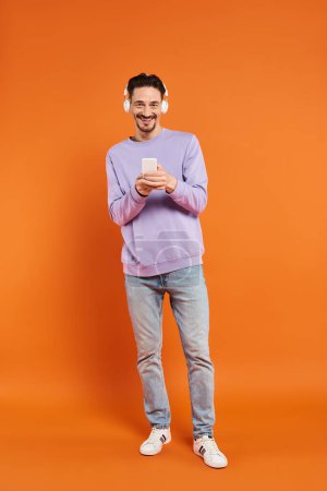 glücklicher Mann mit drahtlosen Kopfhörern, Musik hörend und Smartphone auf orangefarbenem Hintergrund