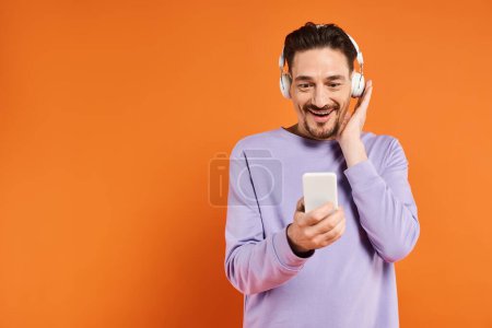 Lächelnder Mann mit drahtlosen Kopfhörern, Musik hörend und Smartphone auf orangefarbenem Hintergrund