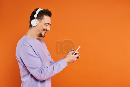 glücklicher Mann in drahtlosen Kopfhörern, der Musik und SMS auf dem Smartphone auf orangefarbenem Hintergrund hört