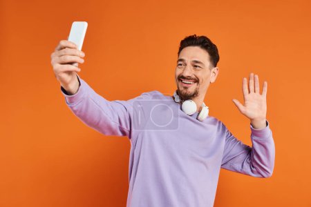positiver Mann mit drahtlosen Kopfhörern winkt bei Videoanruf auf Smartphone auf orangefarbenem Hintergrund