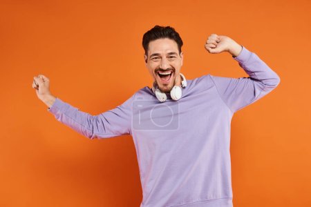 Téléchargez les photos : Homme joyeux en pull violet avec écouteurs sans fil posant sur fond orange, amateur de musique - en image libre de droit