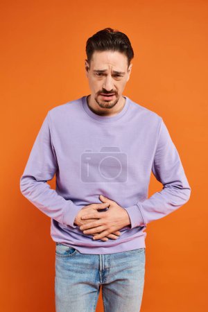 bärtiger Mann in lila Pullover und Jeans mit Bauchschmerzen auf orangefarbenem Hintergrund, Schmerzen