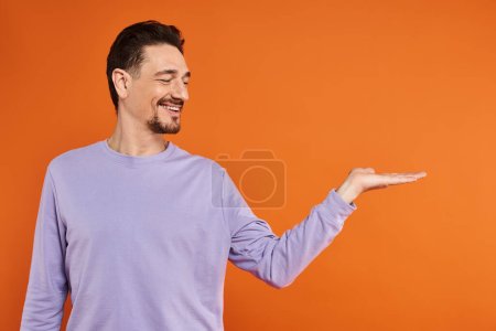 glücklicher bärtiger Mann in lila Sweatshirt präsentiert, während er mit der Hand auf orangefarbenen Hintergrund zeigt