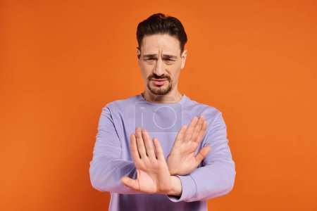 bearded man in purple sweater showing refusal gesture with his hands on orange background
