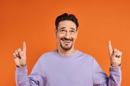 cheerful man in purple sweatshirt pointing up with fingers on orange background, presenting