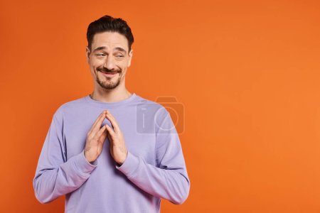 bearded man with hopeful expression and folded hands looking away on orange background, aspiration