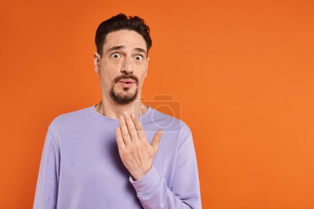 bearded surprised man in purple sweatshirt looking at camera on orange background, astonishment