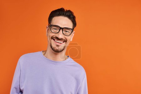 fröhlicher Mann in lila Sweatshirt und trendiger Brille auf orangefarbenem Hintergrund, positive Emotion