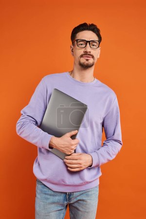 bearded man in eyeglasses and purple sweater holding laptop and standing on orange background