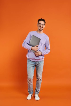 full length of bearded man in eyeglasses and purple sweater holding laptop on orange background