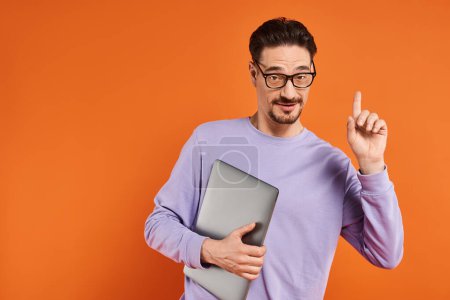 bearded man in eyeglasses and purple sweater holding laptop and pointing up on orange background
