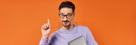 homme barbu dans des lunettes et pull violet tenant ordinateur portable et pointant vers le haut sur fond orange, bannière