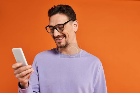 hombre alegre en gafas y suéter púrpura usando teléfono inteligente sobre fondo naranja, mensajes de texto