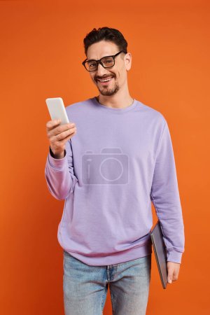 hombre feliz en gafas y suéter púrpura usando teléfono inteligente sobre fondo naranja, mensajes de texto
