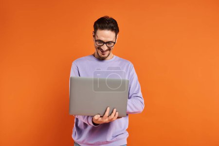 bärtiger Mann mit Brille und lila Pullover mit Laptop auf orangefarbenem Hintergrund, Fernarbeit