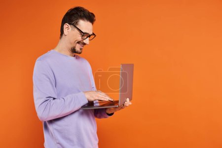 smiling man in eyeglasses and purple sweater using laptop on orange background, remote work