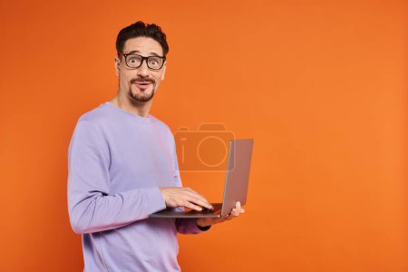 homme étonné dans les lunettes et le pull violet en utilisant un ordinateur portable sur fond orange, travail à distance