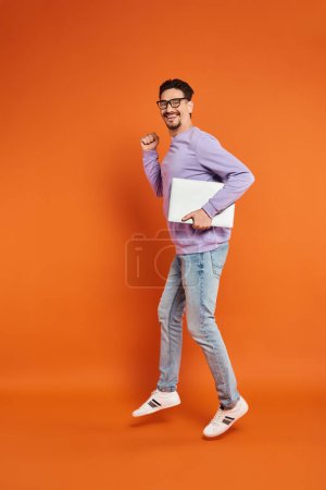 happy bearded man in eyeglasses and purple sweater walking with laptop on orange background