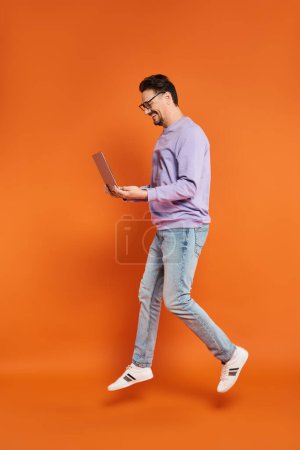 cheerful man in eyeglasses and purple sweater using laptop on orange background, remote work