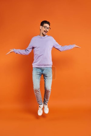 hombre alegre en gafas y suéter púrpura levitando sobre fondo naranja, volando en el aire