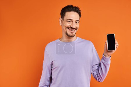 happy man in glasses and purple sweater holding smartphone with blank screen on orange background