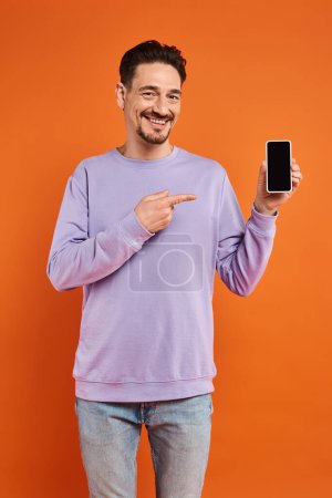 glücklicher Mann mit Brille und lila Pullover zeigt auf Smartphone mit leerem Bildschirm auf orangefarbenem Hintergrund