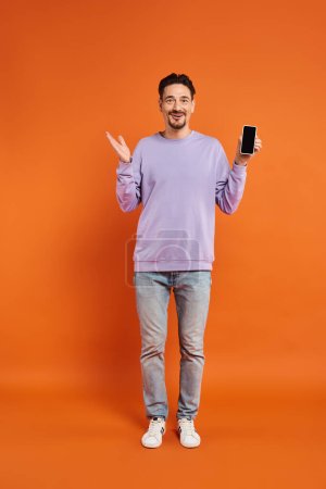amazed man in glasses and purple sweater holding smartphone with blank screen on orange background