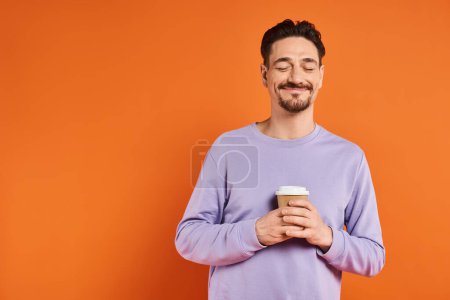 pleased and bearded man holding paper cup with coffee to go on orange background, enjoyment