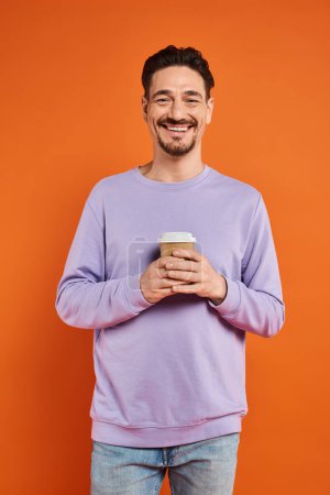 cheerful and bearded man holding paper cup with coffee to go on orange background, enjoyment