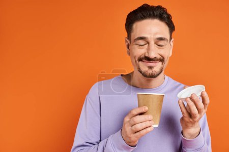 fröhlicher Mann mit Bart hält Pappbecher mit Kaffee auf orangefarbenem Hintergrund, Genuss