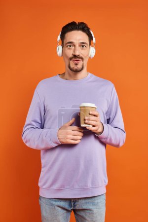 surprised man in wireless headphones holding paper cup with coffee on orange background