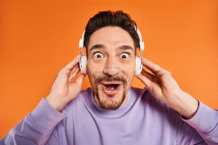 hombre barbudo asombrado en auriculares inalámbricos escuchando música y mirando a la cámara en fondo naranja
