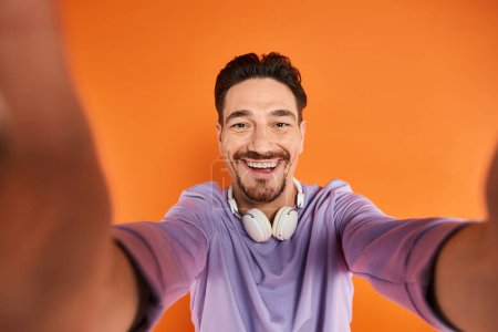 hombre barbudo feliz en auriculares inalámbricos escuchando música y mirando a la cámara en fondo naranja