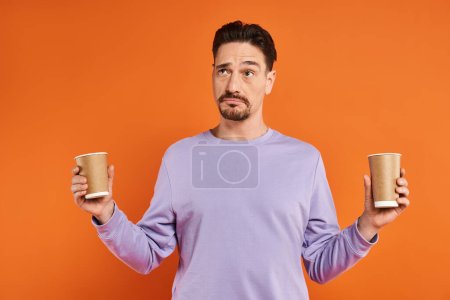 bearded man in purple sweater holding paper cups with coffee to go on orange background, choice