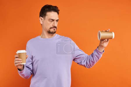 bärtiger Mann im lila Pullover mit leerem Pappbecher auf orangefarbenem Hintergrund, Coffee to go