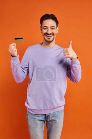 cheerful man with beard shoeing thumb up and holding credit card on orange background, like