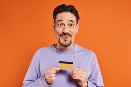 fröhlicher Mann mit Bart lächelt und hält Kreditkarte auf orangefarbenem Hintergrund und blickt in die Kamera