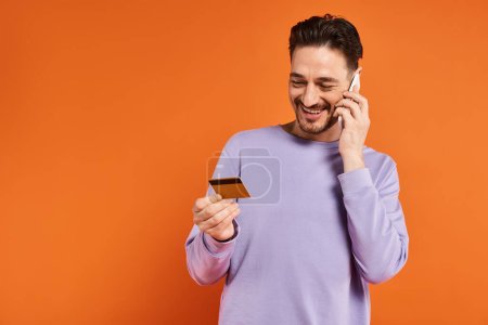 Foto de Hombre alegre sonriendo y sosteniendo la tarjeta de crédito mientras habla en el teléfono inteligente sobre fondo naranja - Imagen libre de derechos