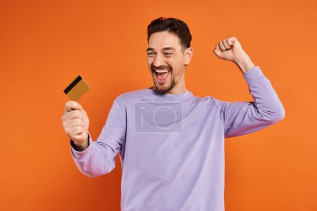 aufgeregter Mann mit Bart lächelt und hält Kreditkarte auf orangefarbenem Hintergrund, jubelnde Geste
