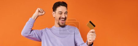 excited man with beard smiling and holding credit card on orange background, rejoicing banner