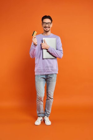 happy man in glasses standing with credit card and laptop on orange background, online shopping