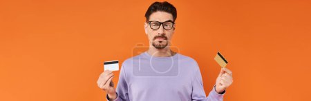 Skeptischer Mann mit Brille und lila Pullover vergleicht zwei Kreditkarten auf orangefarbenem Hintergrund, Banner