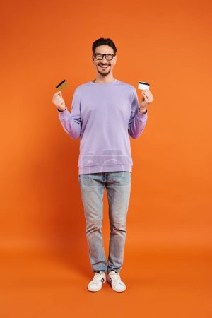happy bearded man in glasses holding credit cards on orange background, shopping and consumerism