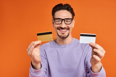 fröhlicher bärtiger Mann in Brille mit Kreditkarten auf orangefarbenem Hintergrund, Einkaufen und Konsum