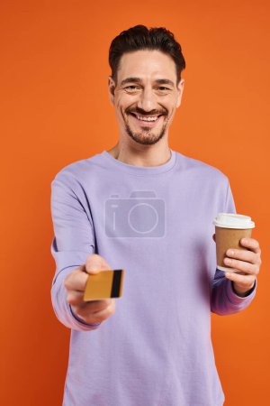 Foto de Hombre feliz en suéter púrpura sosteniendo café para ir y ofreciendo tarjeta de crédito sobre fondo naranja - Imagen libre de derechos