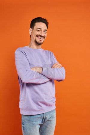 happy man in purple sweater and jeans standing with crossed arms on orange background, casual wear