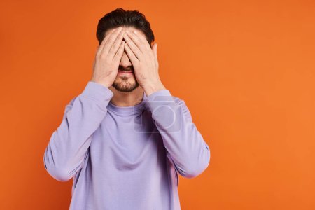 man with beard in purple casual sweater hiding face with hands on orange background, playful mood